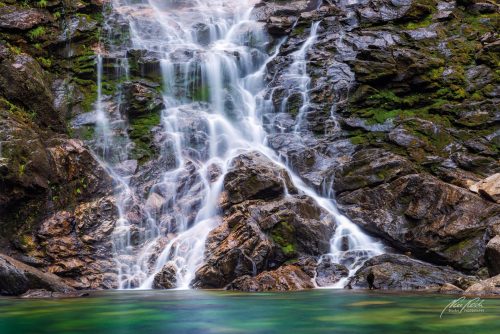 LBK707-8434-Verspielter-kleiner-Wasserfall-ueber-tuerkisgruenem-Bergsee